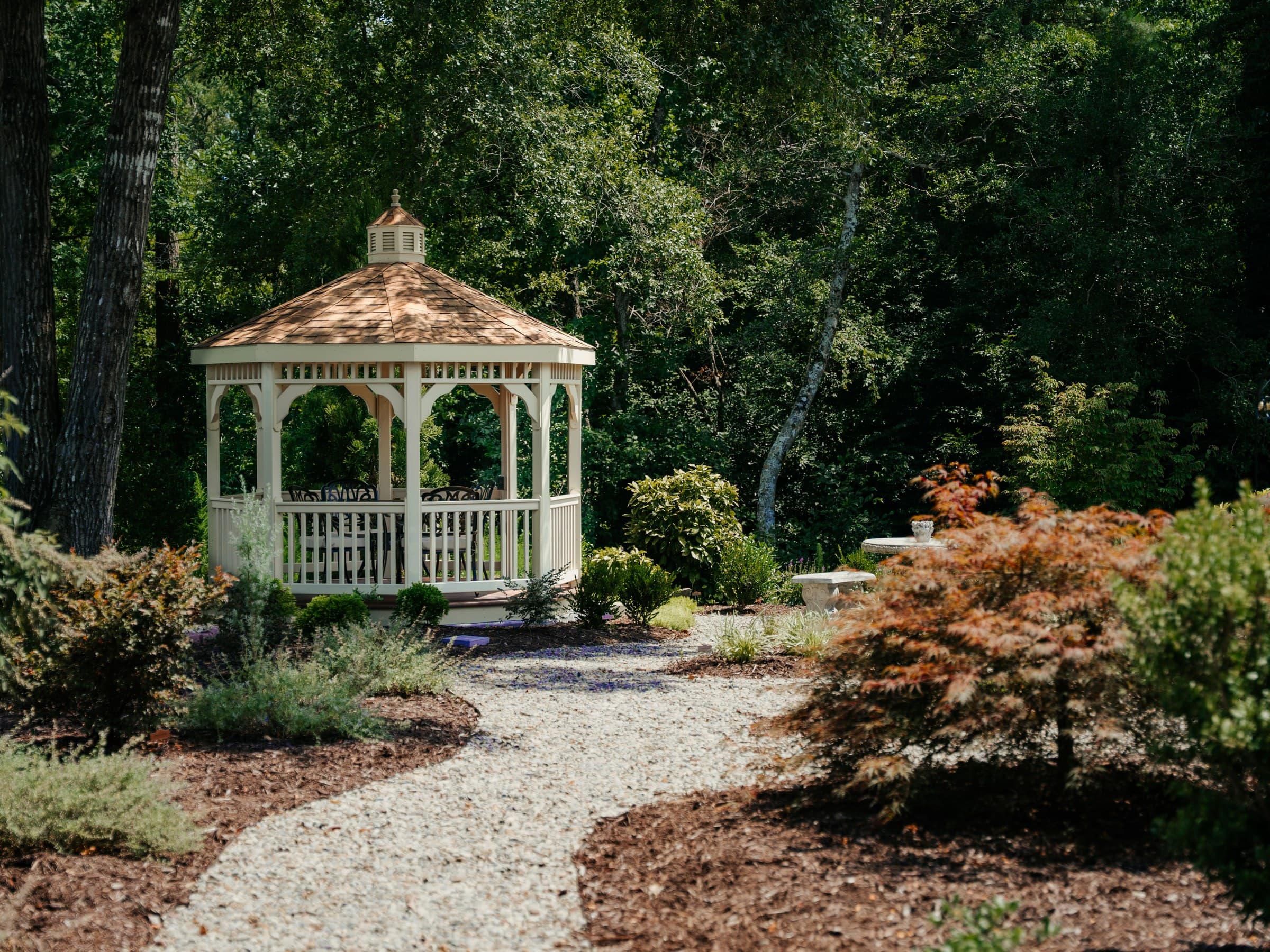 A cedar-roofed garden gazebo at the end of a stone path, framed by Japanese maples and layered conifer beds.
